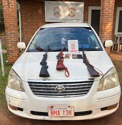 Automóvil blanco con armas sobre el capó, frente a edificio de investigación criminal de la policía.