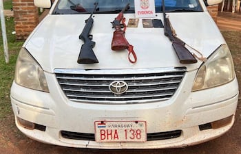 Automóvil blanco con armas sobre el capó, frente a edificio de investigación criminal de la policía.