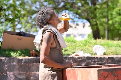 Hombre desaliñado con camiseta sin mangas y trapo en el cuello, bebiendo de una botella en un entorno urbano.