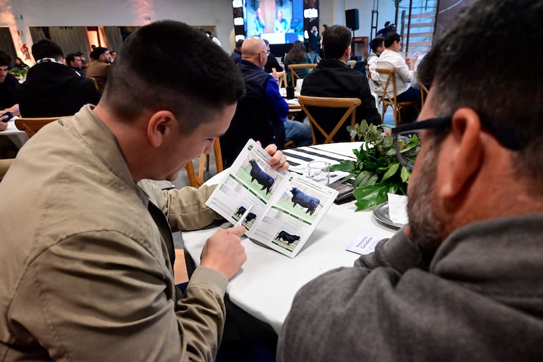 Hombres en chaquetas revisan folleto sobre toros en un evento con público de fondo y decoración sencilla.