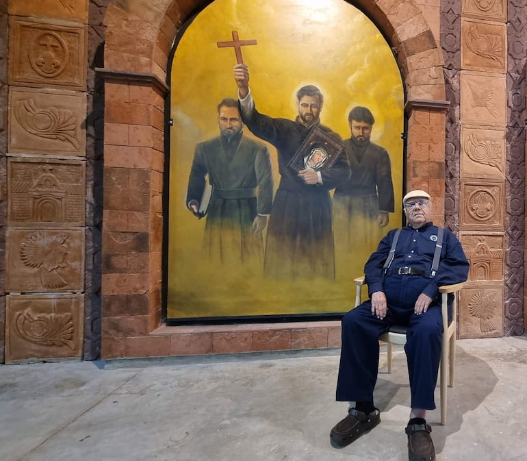Manuel de Jesús Viedma Romero, con camisa azul y sombrero claro, sentado frente a un mural con figuras religiosas.