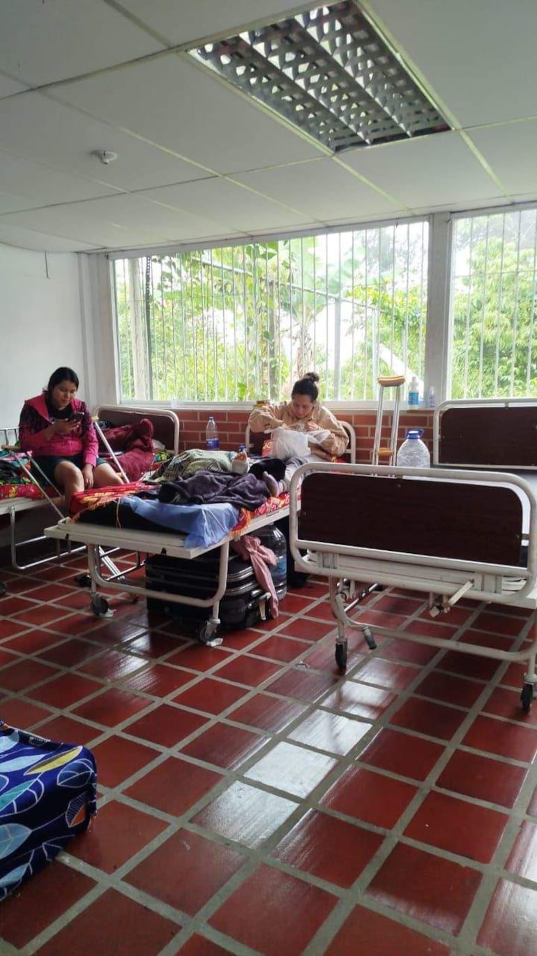 Dos mujeres en ambiente hospitalario: una sentada con teléfono y otra comiendo, cerca de camas cargadas con pertenencias.