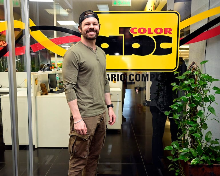 Blake Webb, vestido con camiseta de manga larga y pantalones cargo, sonríe frente a una puerta de vidrio del diario ABC Color.