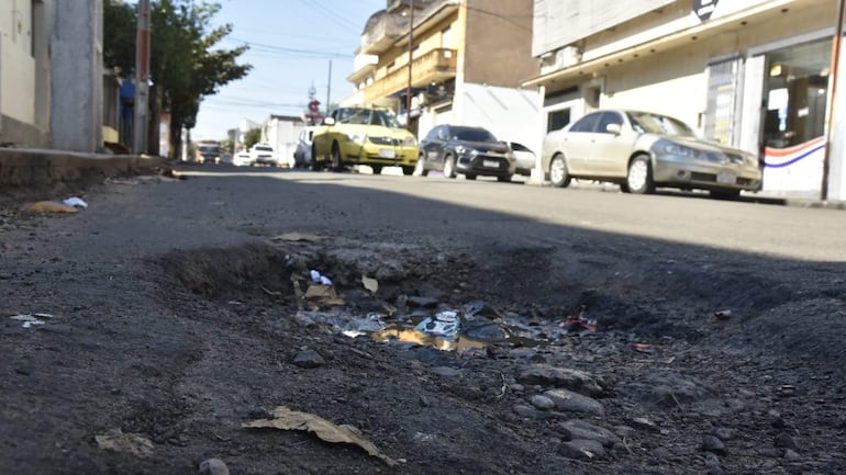 Bache en calle asuncena con agua estancada y basura; edificios comerciales y vehículos pasan por los lados.