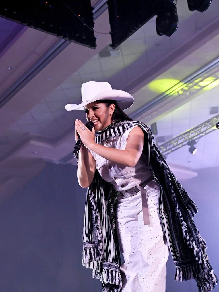 Mujer en vestido blanco brillante y sombrero, sosteniendo un micrófono con manos en gesto de agradecimiento en un escenario.