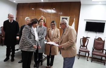 Mirtha Arias en atuendo claro, sosteniendo un documento; rodeada de mujeres jóvenes vestidas formalmente en un ambiente serio.