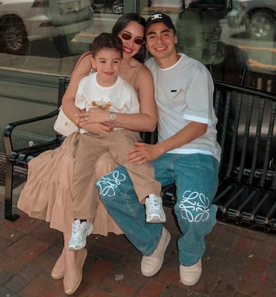 Miguel Almirón y Alexia Notto sonríen mientras abrazan a su hijo Francesco en un banco, rodeados de un ambiente urbano.