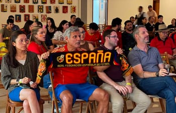 Hombre canoso con camiseta roja y bufanda que dice 'ESPAÑA', mujer con gafas en ambiente festivo de un bar lleno de fanáticos.