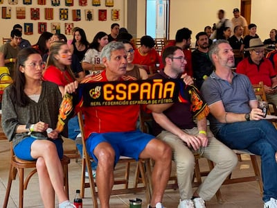 Hombre canoso con camiseta roja y bufanda que dice 'ESPAÑA', mujer con gafas en ambiente festivo de un bar lleno de fanáticos.
