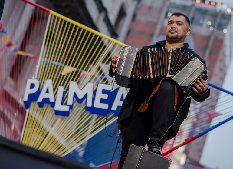 Músico robusto con barba toca acordeón sobre tarima, vistiendo camiseta oscura, fondo decorado con inscripción 'PALMEA'.