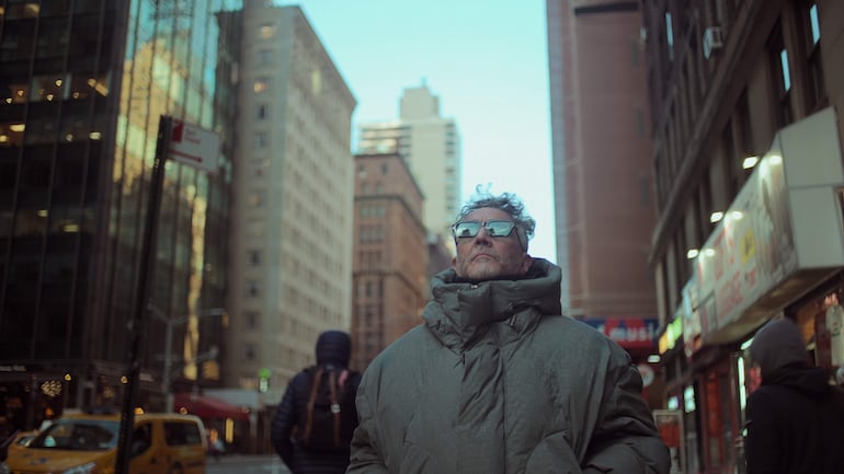 Fito Páez en abrigo voluminoso y gafas de sol, observando el cielo en una calle con edificios altos alrededor.