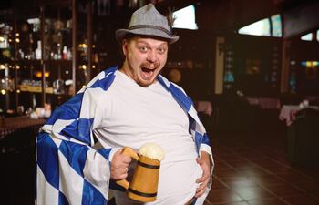 Un hombre con la bandera de Baviera a manera de capa se agarra el estómago con una mano, mientras con la otra sostiene una falsa manija de chopp con cerveza.