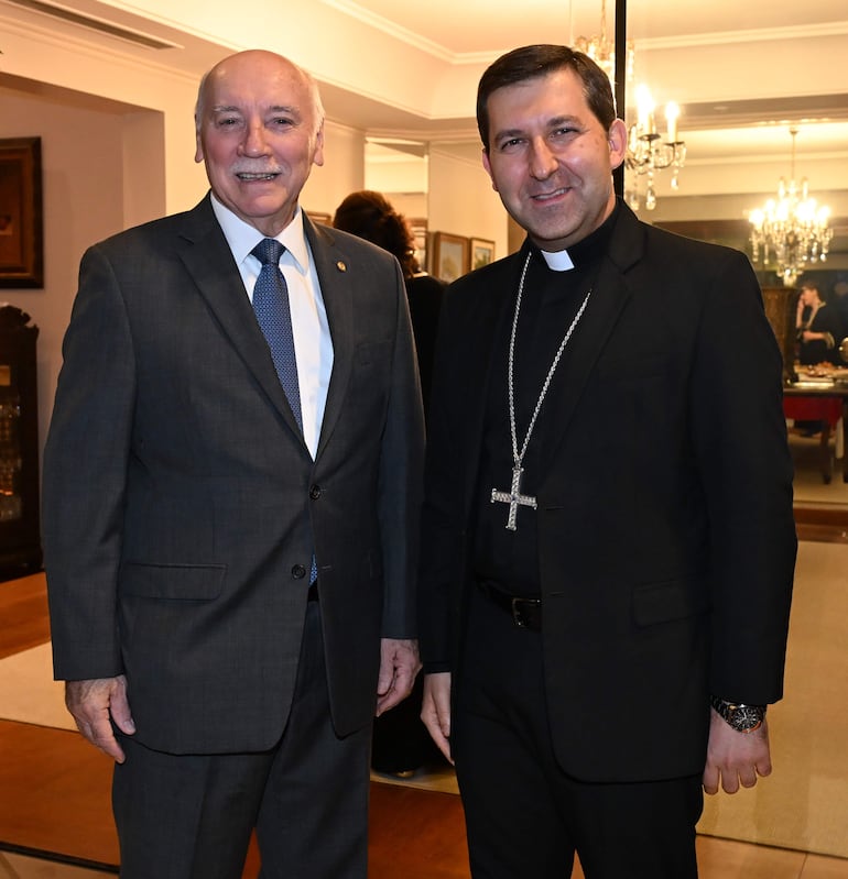 Vincenzo Turturro en traje gris y Eladio Loizaga en hábito religioso, sonriendo en un elegante salón decorado con candelabros.