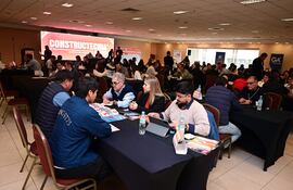 Cuatro personas en mesa: dos hombres revisan materiales, mientras dos mujeres toman notas y observan en un ambiente de feria.