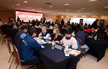 Cuatro personas en mesa: dos hombres revisan materiales, mientras dos mujeres toman notas y observan en un ambiente de feria.