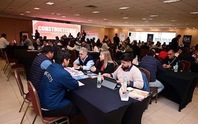 Cuatro personas en mesa: dos hombres revisan materiales, mientras dos mujeres toman notas y observan en un ambiente de feria.