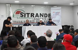 Hombres en mesa de diálogo; uno habla con chaleco negro, otros escuchan atentos. Fondo con bandera del sindicato.