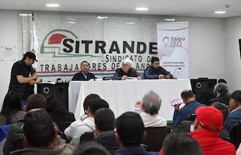 Hombres en mesa de diálogo; uno habla con chaleco negro, otros escuchan atentos. Fondo con bandera del sindicato.