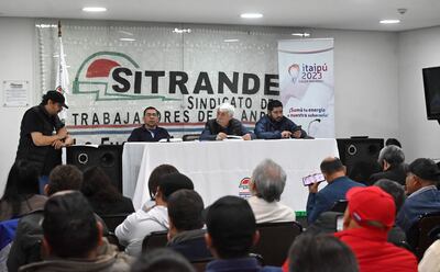 Hombres en mesa de diálogo; uno habla con chaleco negro, otros escuchan atentos. Fondo con bandera del sindicato.