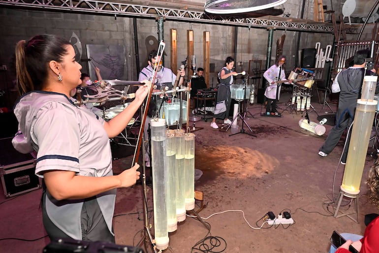 Músicos en atuendos grises interpretan música en un ambiente con paredes de ladrillo expuesto y suelo húmedo.