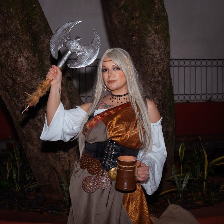 Mujer con cabello rubio y disfraz medieval, blusa blanca y falda dorada, sostiene un hacha y un recipiente en parque.