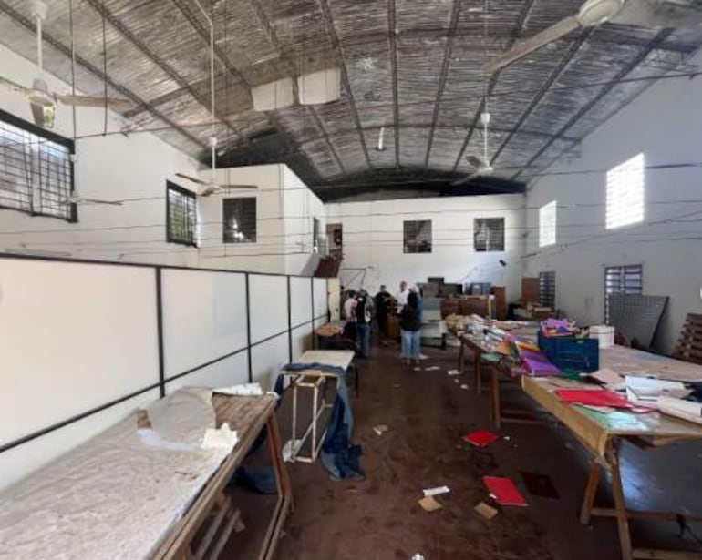 Seis personas en un salón iluminado trabajan sobre mesas desordenadas, con hojas de papel y objetos variados alrededor.