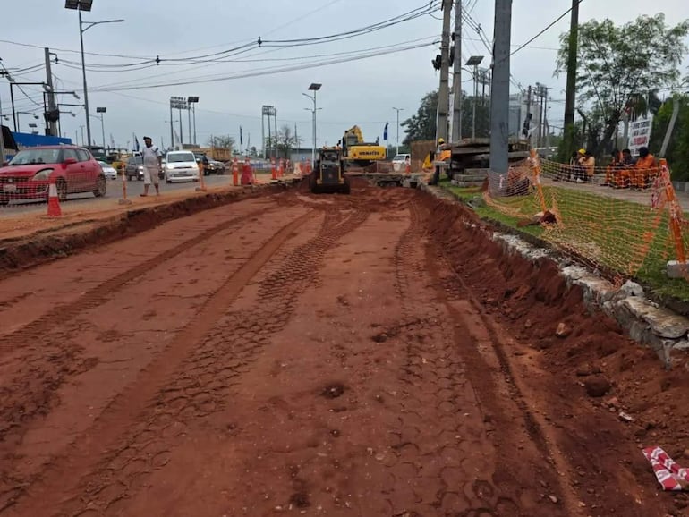 Trabajador en ropa clara supervisa obras viales en terreno de tierra roja, con maquinaria amarilla en acción y autos alrededor.