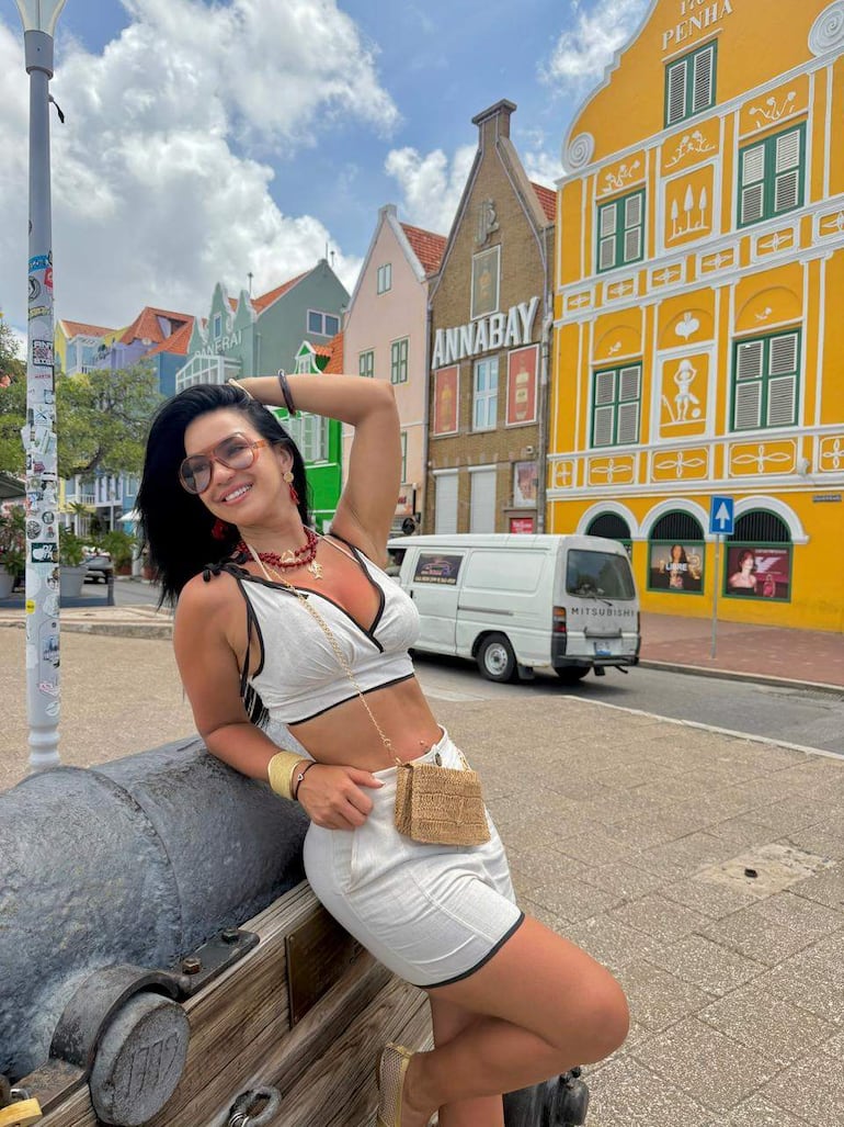 Mujer con blusa de tirantes y falda blanca, sentada sobre un cañón, sonriente, con fondo arquitectónico colorido.