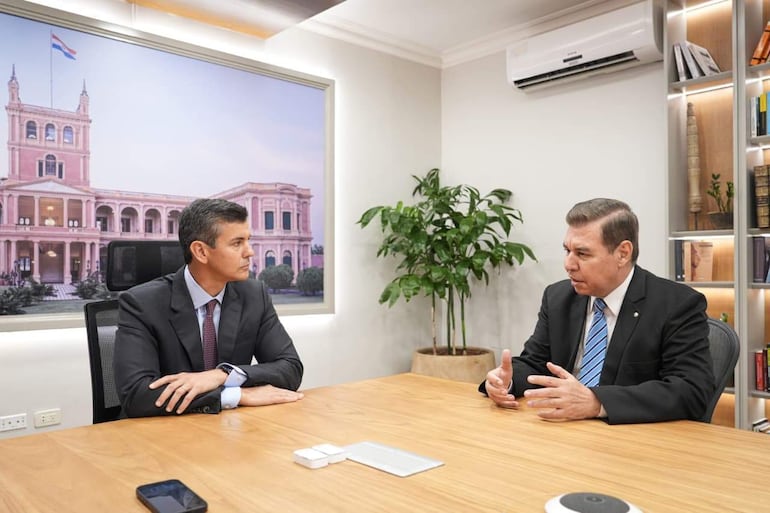 Jacinto Santa María en traje gris escuchando a Santiago Peña Palacios, en traje negro y corbata a rayas, en un entorno de reunión formal.