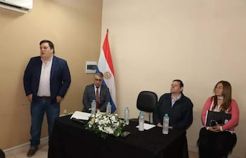 César Sosa de pie en chaqueta oscura, Francisco Ruiz Díaz con gafas escuchando, y otros participantes sentados en sala con bandera paraguaya.