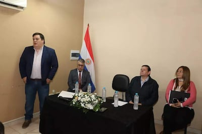 César Sosa de pie en chaqueta oscura, Francisco Ruiz Díaz con gafas escuchando, y otros participantes sentados en sala con bandera paraguaya.
