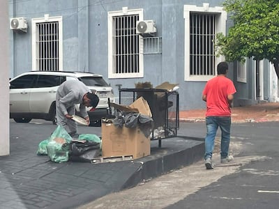 Dos hombres en una calle de Asunción, uno revisa un basurero mientras el otro camina, rodeados de basura y edificios.