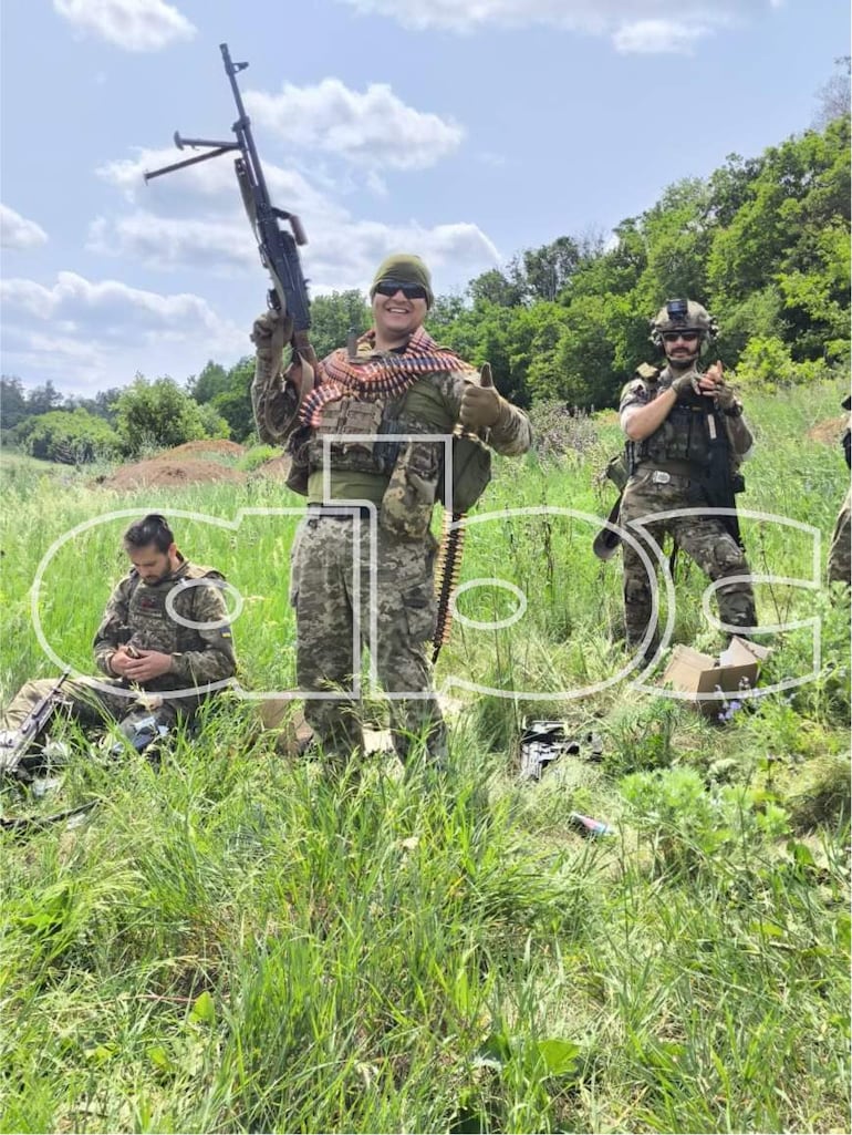 Eduardo Román con rifle, sonriente y pulgar arriba, rodeado de tres soldados en uniforme militar en un paisaje natural.