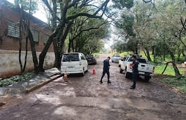 Hombre vestido de negro en movimiento junto a un oficial de policía cerca de vehículos en una calle de tierra arbolada.