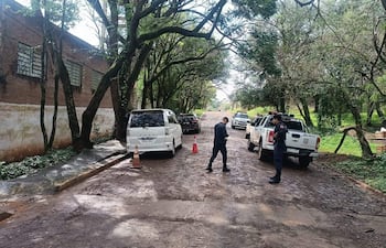 Hombre vestido de negro en movimiento junto a un oficial de policía cerca de vehículos en una calle de tierra arbolada.