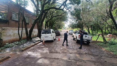 Hombre vestido de negro en movimiento junto a un oficial de policía cerca de vehículos en una calle de tierra arbolada.