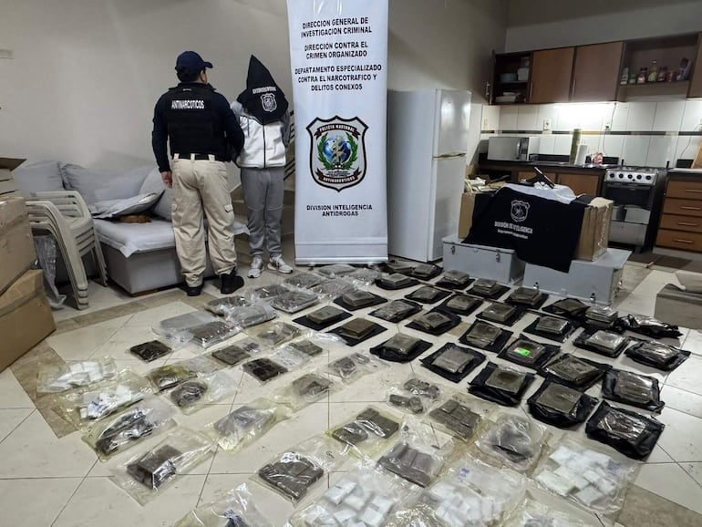 Agentes policiales con chalecos 'ANTINARCÓTICOS', rodeados de paquetes de drogas en ambiente de vivienda.