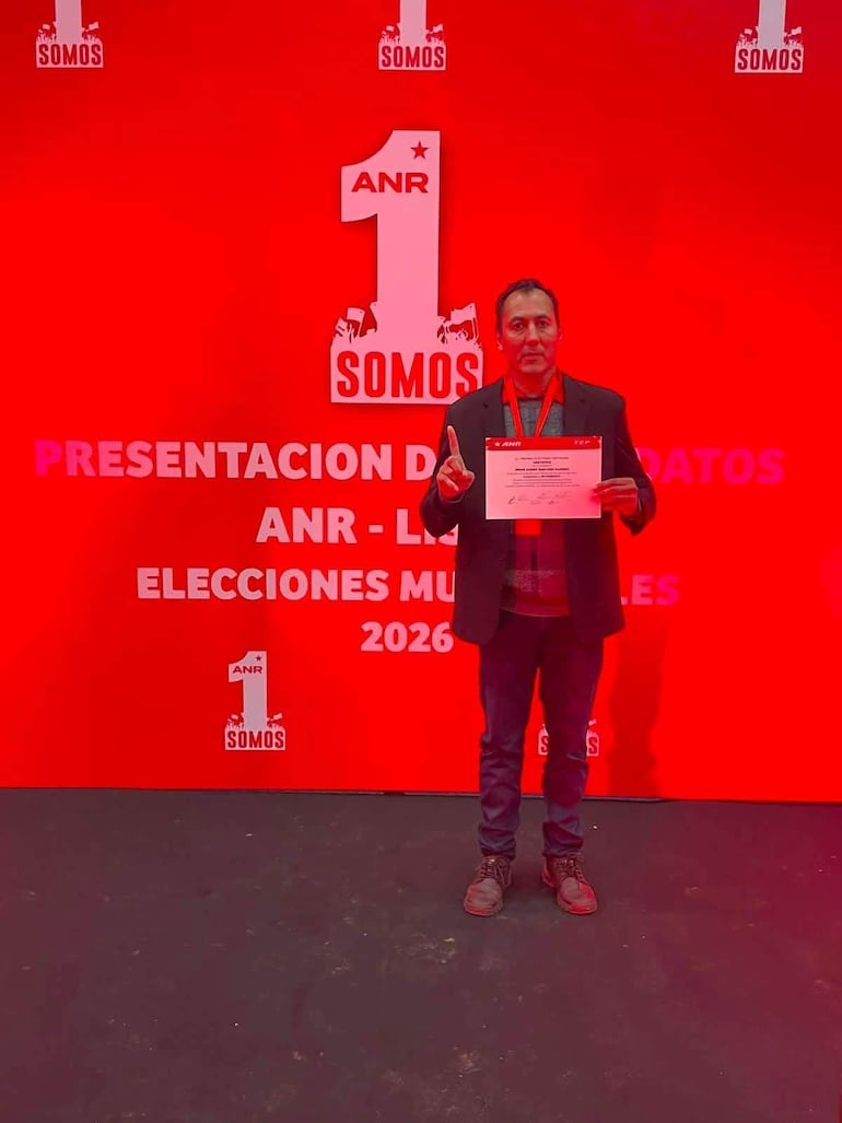Hombre de pie en traje oscuro y camisa de cuadros, sostiene documento y hace gesto de 'número uno' en evento de la ANR.