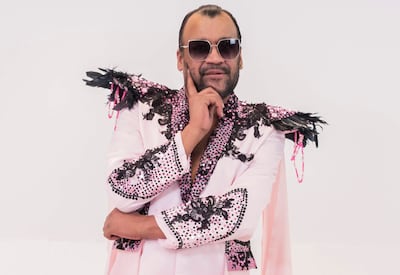 Gustavo Cabaña en traje rosa con detalles negros y gafas de sol, de pie con una mano en el mentón, en un fondo sencillo.