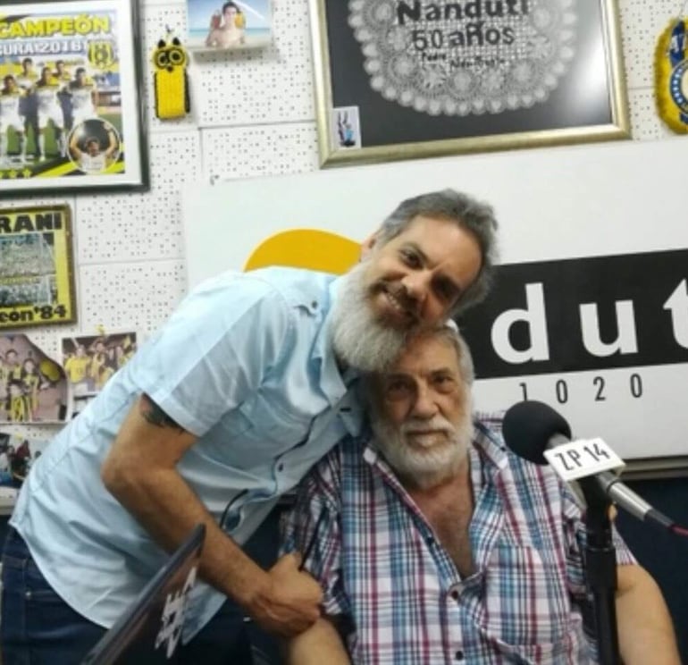 Hermes Leonardo Rubin Leo abraza a Pedro RANI, ambos con barba, en un estudio decorado con recuerdos del medio Nandutí.