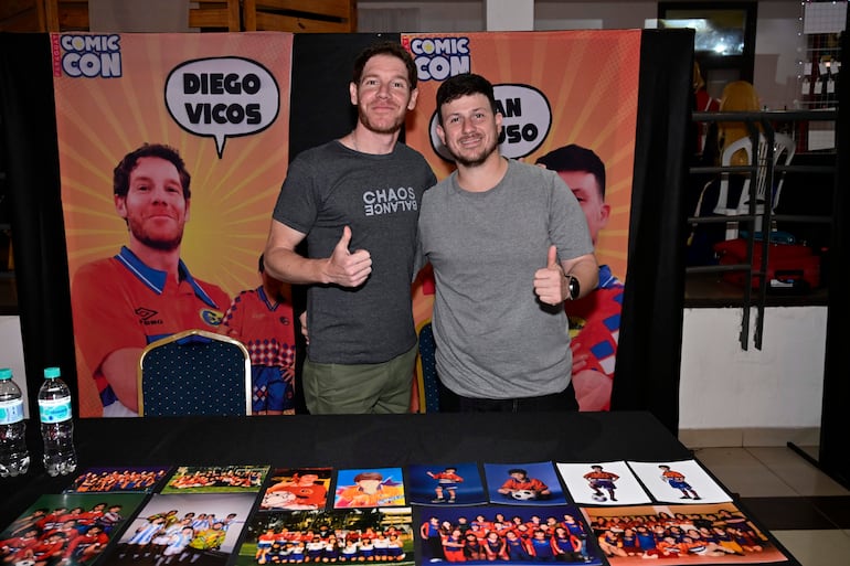 Hombre con camiseta negra y otro con camiseta gris sonríen y levantan el pulgar en un evento colorido de Comic Con.