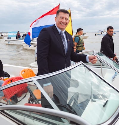 Hombre en traje oscuro sonríe en barco con banderas de Paraguay y otra amarilla, rodeado de varias personas en un día nublado.