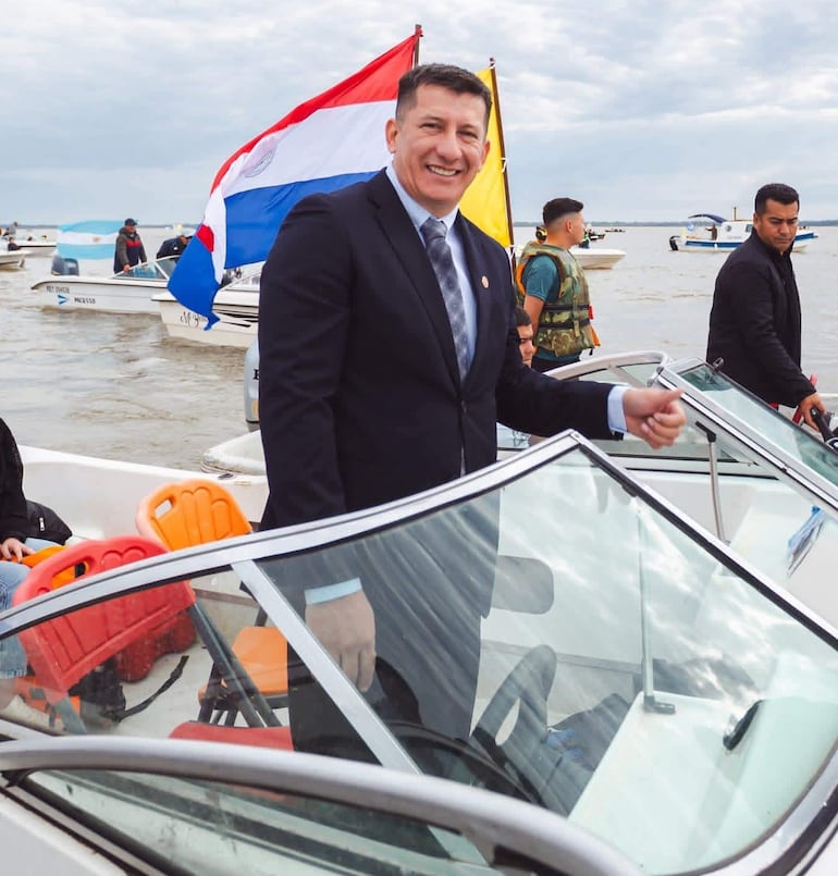 Hombre en traje oscuro sonríe en barco con banderas de Paraguay y otra amarilla, rodeado de varias personas en un día nublado.