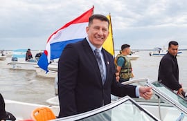 Hombre en traje oscuro sonríe en barco con banderas de Paraguay y otra amarilla, rodeado de varias personas en un día nublado.