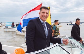 Hombre en traje oscuro sonríe en barco con banderas de Paraguay y otra amarilla, rodeado de varias personas en un día nublado.