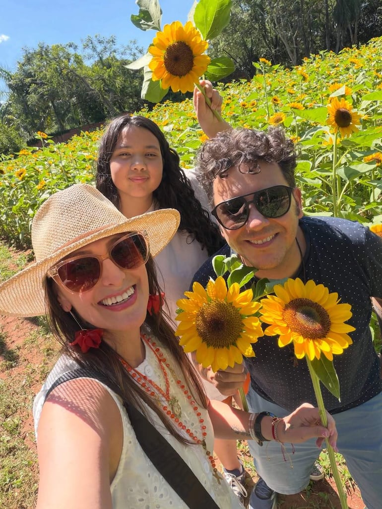 Mujer con blusa blanca y sombrero de paja sonríe junto a una niña y un hombre con gafas de sol, todos en un campo de girasoles.