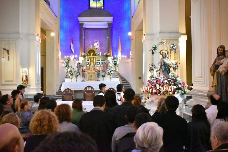 Asistentes de pie en ceremonia religiosa, algunos con teléfonos en mano, mientras dos hombres en traje se destacan al frente.