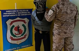 Hombre de espaldas con saco oscuro y capucha, junto a un oficial de policía en uniforme camuflado, en ambiente interior amarillo.