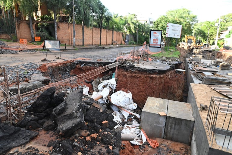 Zona de construcción con zanja abierta, escombros y excavadora, en un ambiente urbano con árboles y muro de ladrillo al fondo.