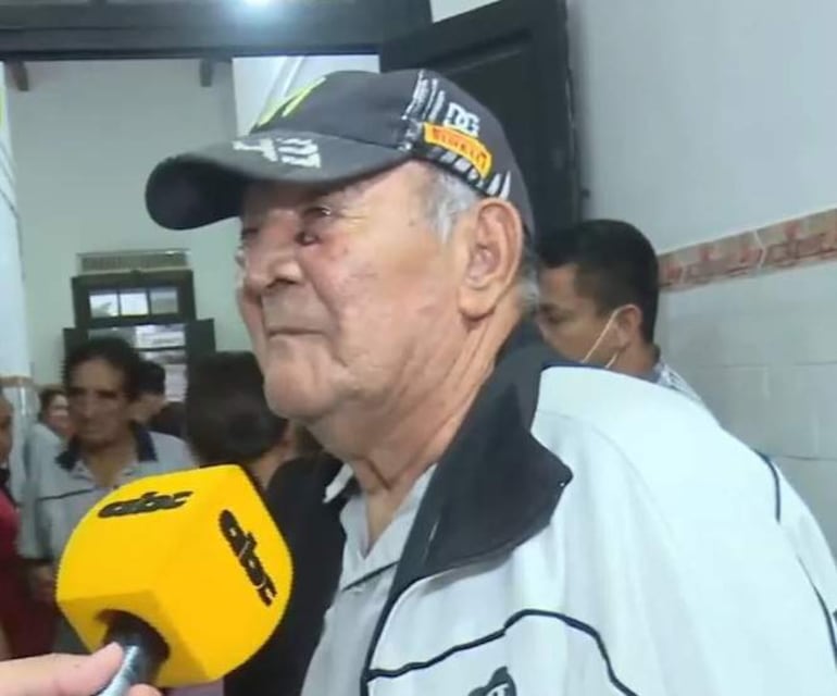 Hombre mayor con chaqueta blanca y visera negra siendo entrevistado en un ambiente público.